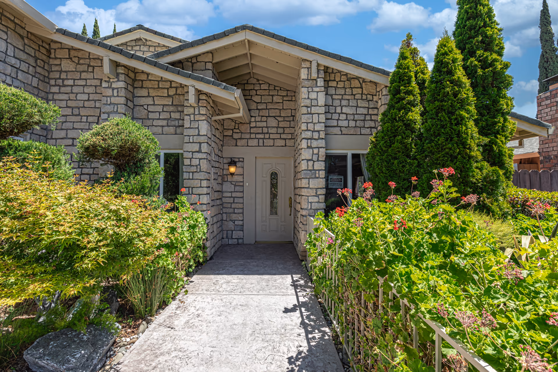 Front entrance of a stone building with a covered porch, surrounded by lush green bushes, flowering plants, and tall evergreen trees under a blue sky with some clouds.