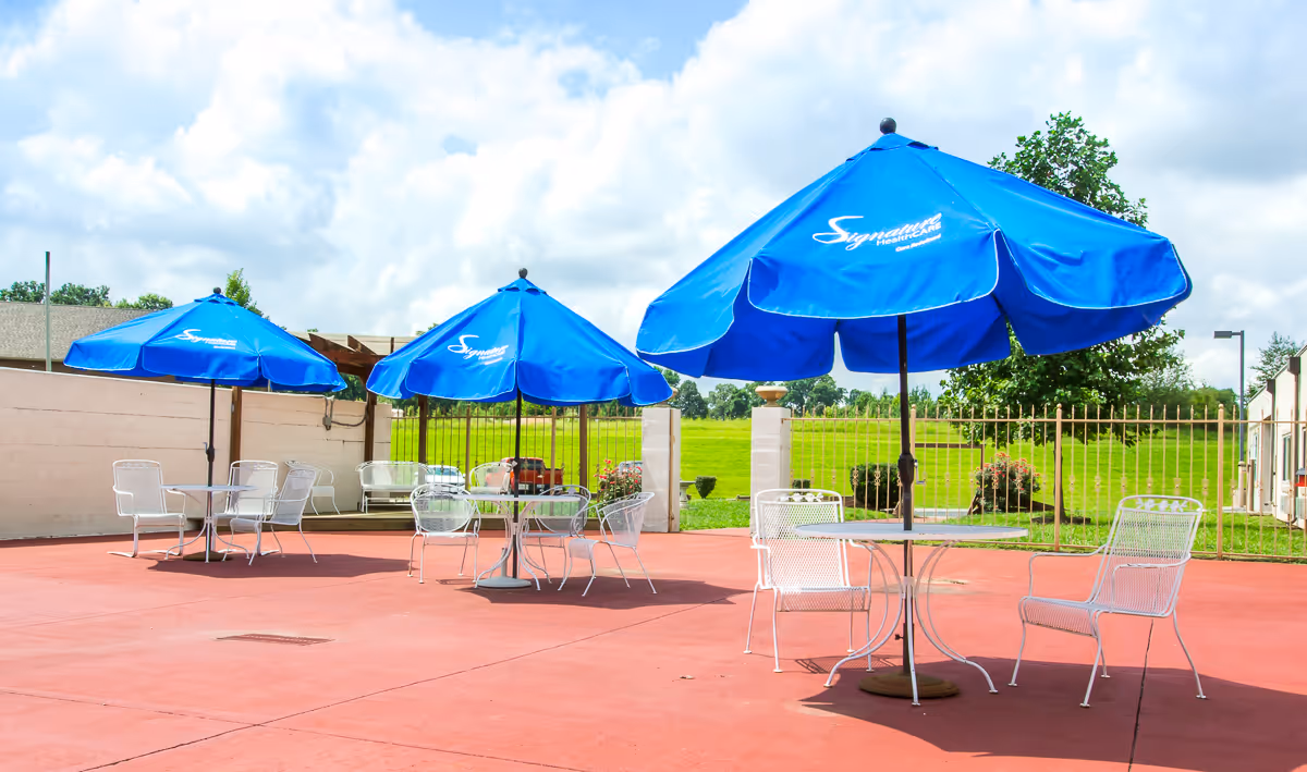 Outdoor patio area with three blue umbrellas labeled Signature HealthCARE, each shading a metal table and chairs. The patio has a red concrete floor and is enclosed by a metal fence with green grass and trees visible beyond.