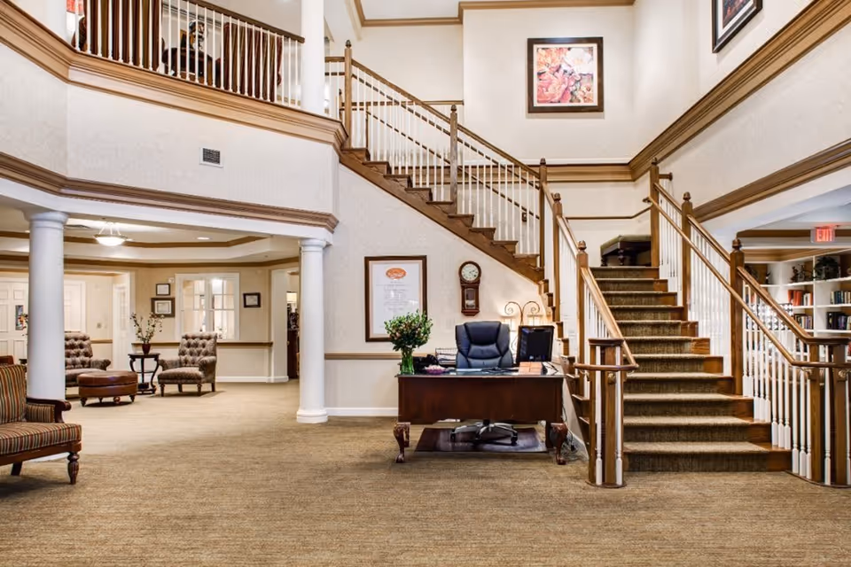 Interior view of a senior living facility lobby with a wooden staircase, a desk with a chair and computer, comfortable seating areas with upholstered chairs and a small table, decorative columns, and framed artwork on the walls.