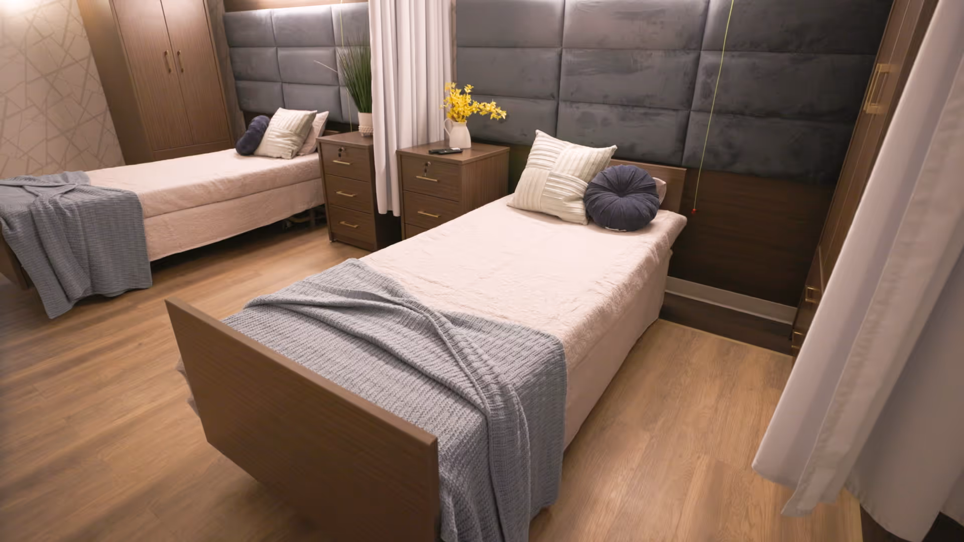 A cozy bedroom with two single beds separated by a wooden nightstand. Each bed has a beige bedspread, a gray throw blanket, and decorative pillows. The room features wooden flooring, a padded headboard, a wardrobe, and a vase with yellow flowers on the nightstand. Light curtains partially frame the scene.