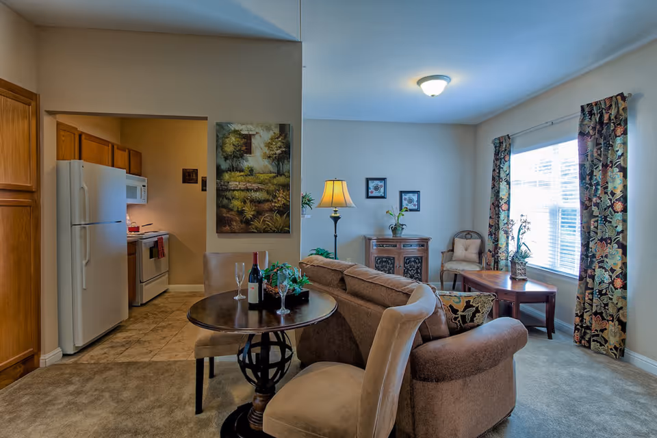 Cozy living room with a sofa, small round dining table, lamp and an open view into a kitchen with a refrigerator; a window with patterned curtains brightens the room.
