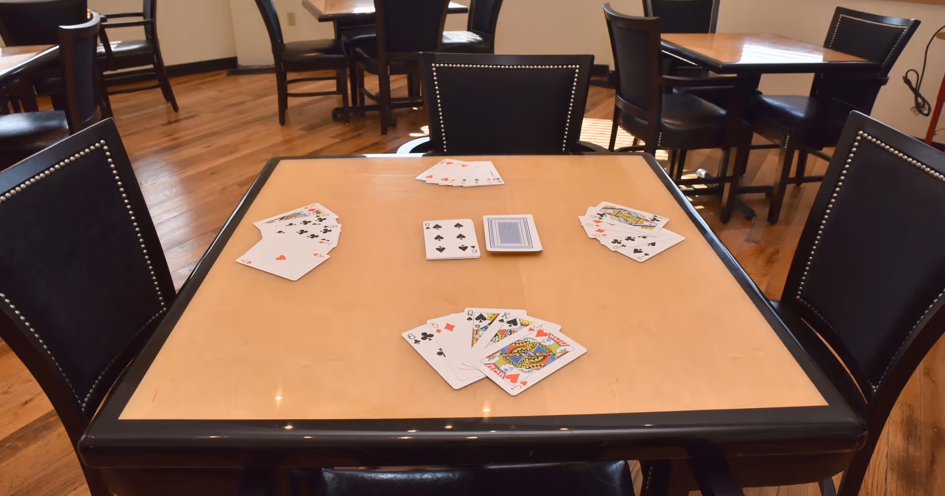 A card game set up on a square table in a communal dining room with black chairs and hardwood floors.