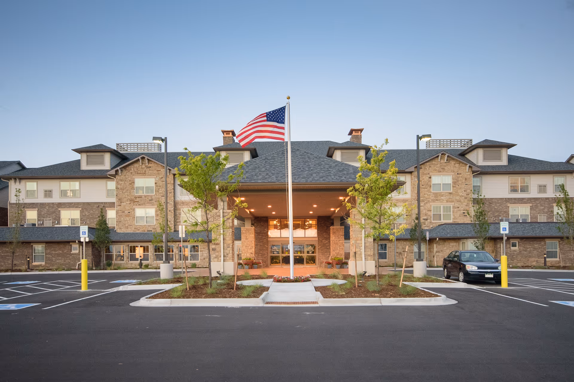 Front entrance of a three-story brick senior living building with an American flag on a pole and a parking lot in the foreground.