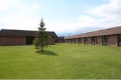 Grassy central courtyard with a single pine tree surrounded by low brick single-story buildings under a partly cloudy sky.
