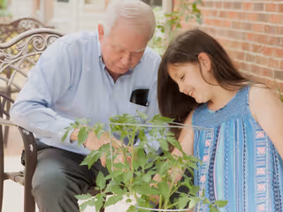 An elderly man and a young girl are sitting together outdoors on a bench near a brick wall, smiling and looking at a plant in front of them.