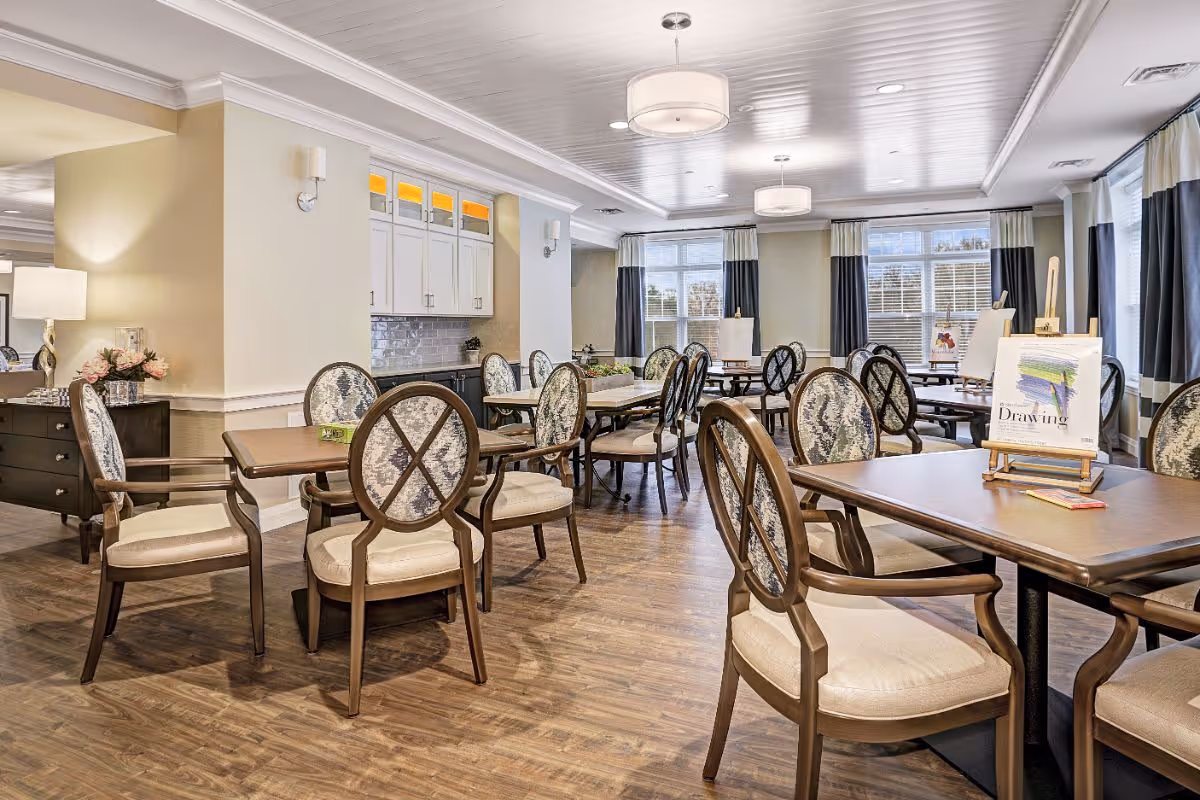 A bright and spacious activity room with multiple wooden tables and cushioned chairs arranged neatly. The room features large windows with curtains, overhead round light fixtures, and a cabinet area with white upper cabinets and a gray tiled backsplash. Some tables have easels with drawing materials displayed on them.
