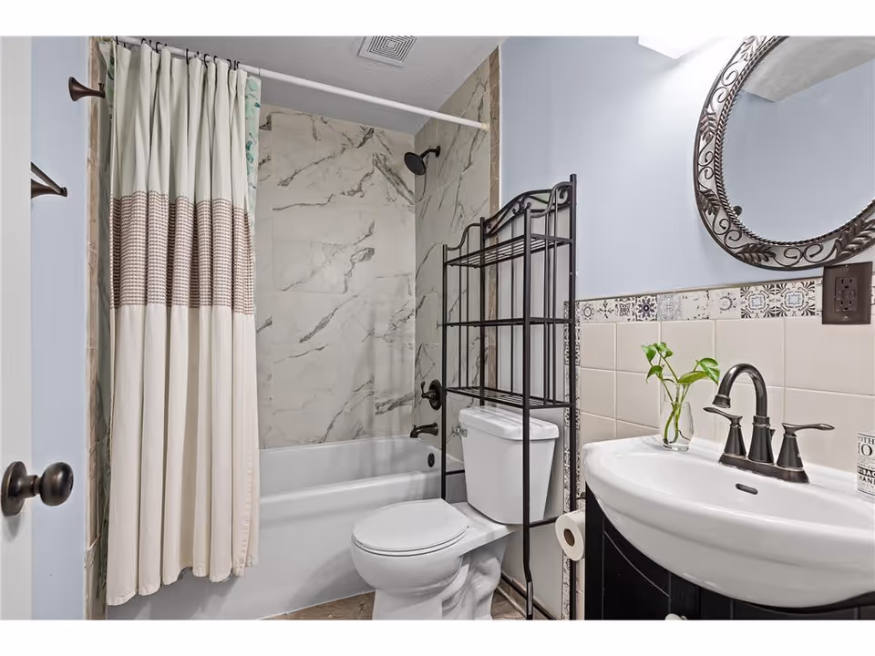 A clean bathroom featuring a white bathtub with a marble-patterned wall surround and a beige and brown striped shower curtain. There is a white toilet with a black metal shelving unit above it. A white sink with a black faucet is next to the toilet, accompanied by a small green plant in a glass vase and a round decorative mirror with a black ornate frame on the wall above the sink.