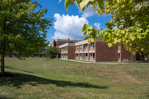 Exterior view of a two-story brick building with multiple windows, surrounded by green grass and trees under a partly cloudy blue sky.