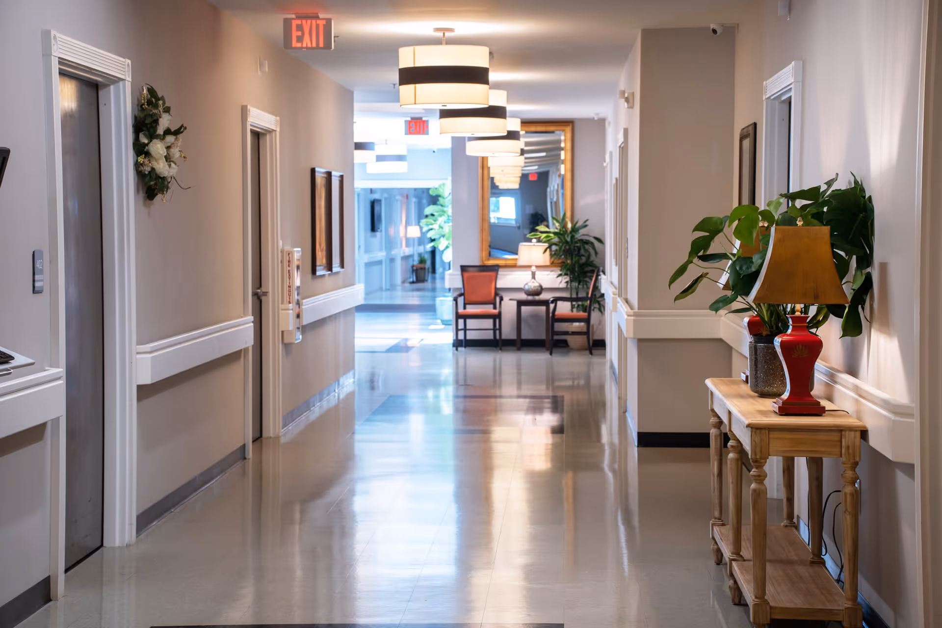 A clean, well-lit hallway in an assisted living facility with beige walls, several closed doors, a wooden table with a red lamp and plants on the right, and chairs with a table and lamp at the far end near a large mirror. Ceiling lights and exit signs are visible.