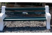 A green wooden bench with white concrete ends reading "Bethany Village" set on a gravel surface.