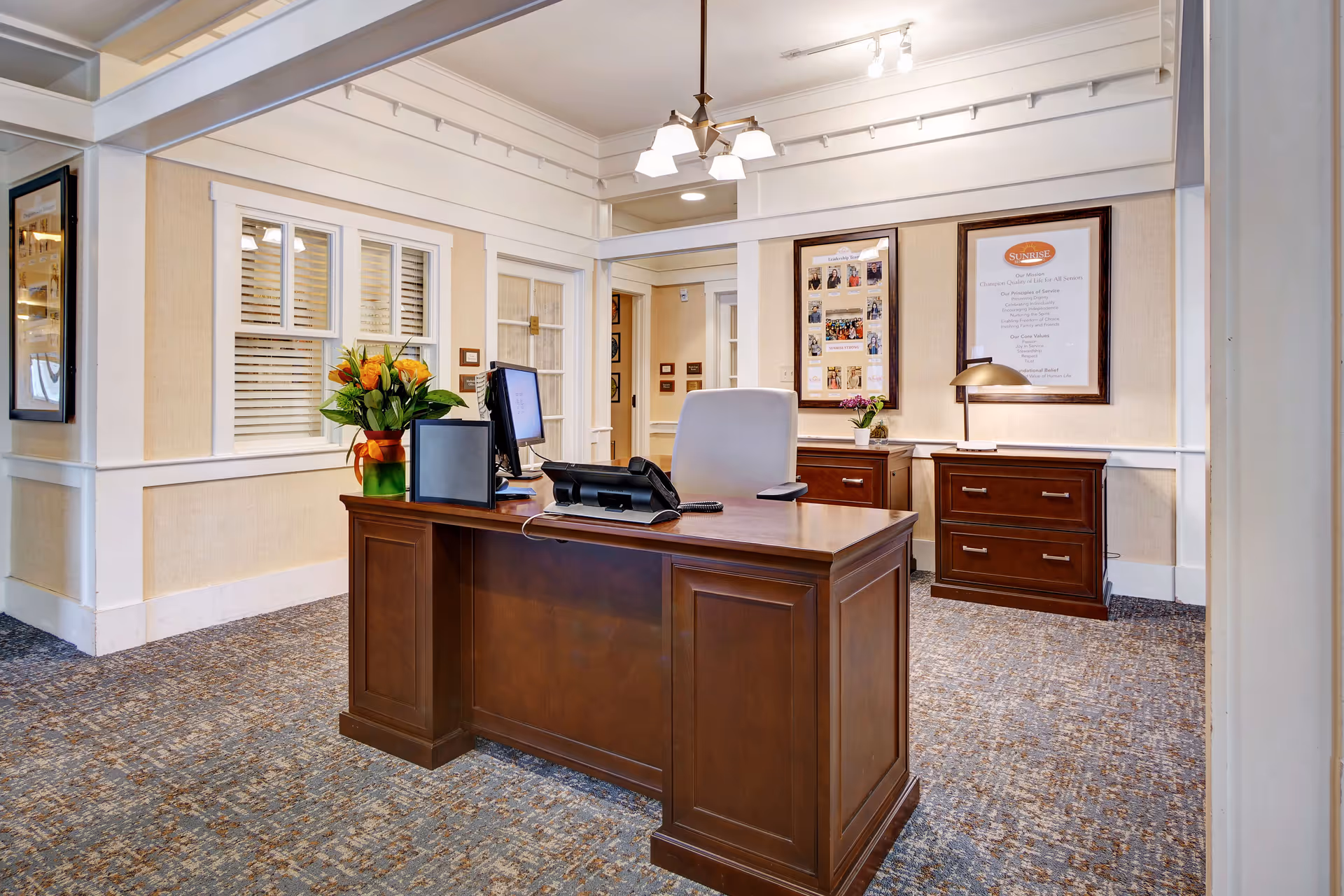 A reception or office area with a wooden desk, computer, phone, and a chair. Behind the desk are two wooden filing cabinets with framed pictures and documents on the wall. The room has beige walls, carpeted floor, and a ceiling light fixture.
