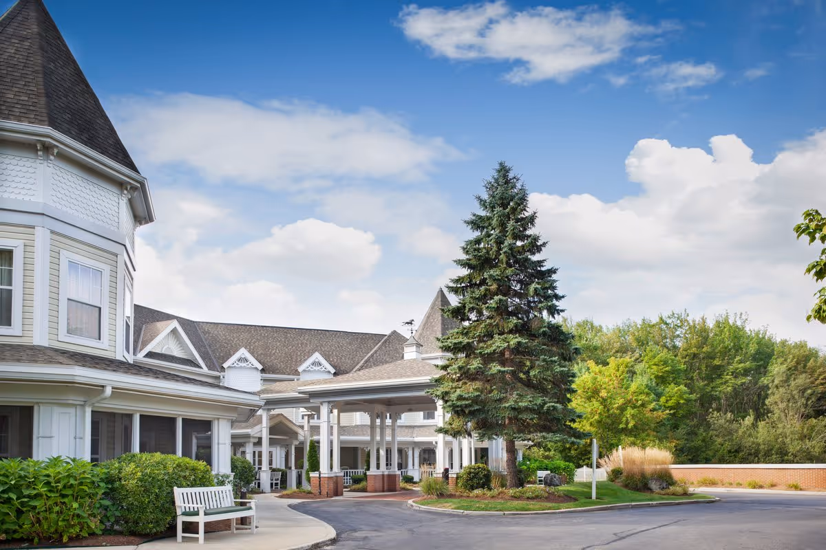 Front exterior of a senior living facility with a covered entrance, driveway, benches, landscaping, and a large evergreen under a blue sky.