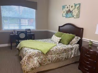 A neatly arranged bedroom with a bed featuring floral patterned bedding and green pillows. There is a wooden nightstand with a lamp on the right side of the bed. A desk and chair are positioned in front of a window with blinds, and a floral wall art hangs above the bed's headboard.