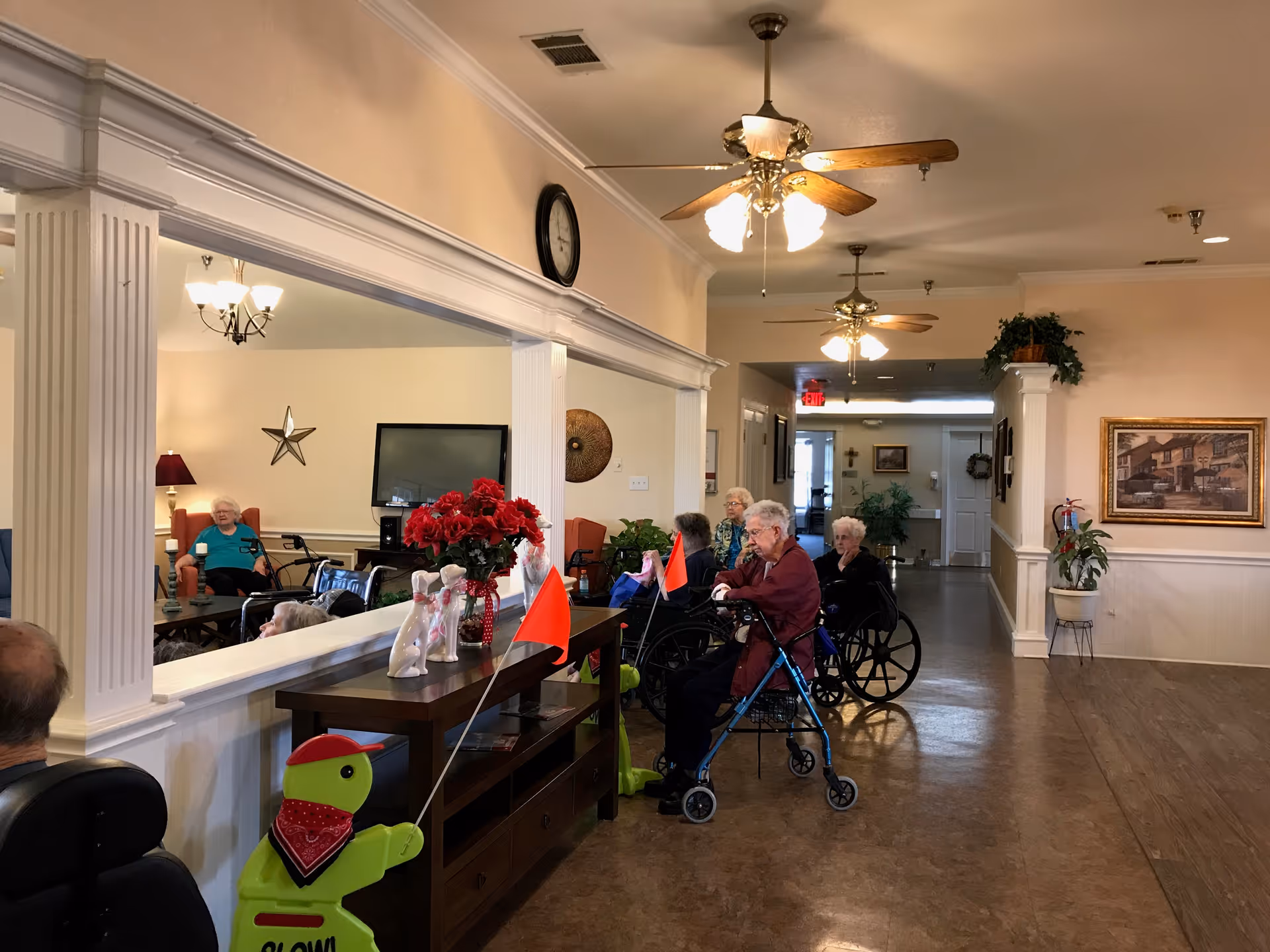 Several elderly residents seated in a communal lounge area with ceiling fans, a console table with flowers, and wheelchairs.