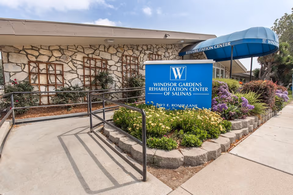 Exterior view of Windsor Gardens Rehabilitation Center of Salinas with blue sign, ramp, and landscaping.