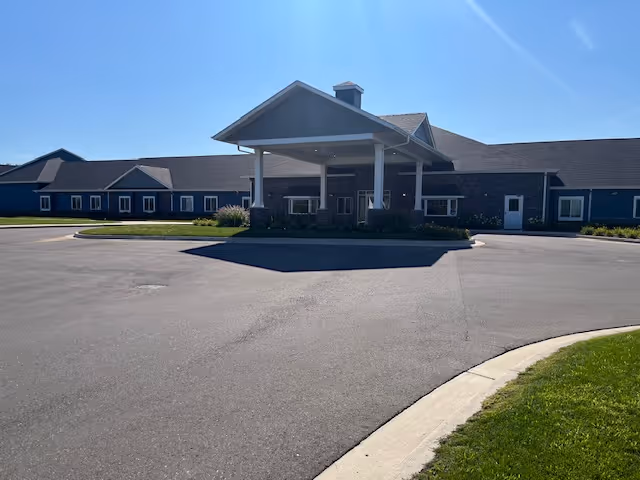Front exterior view of Hampton Manor of Trenton Premier Assisted Living Facility showing a large covered entrance with a driveway and a long building with multiple windows under a clear blue sky.
