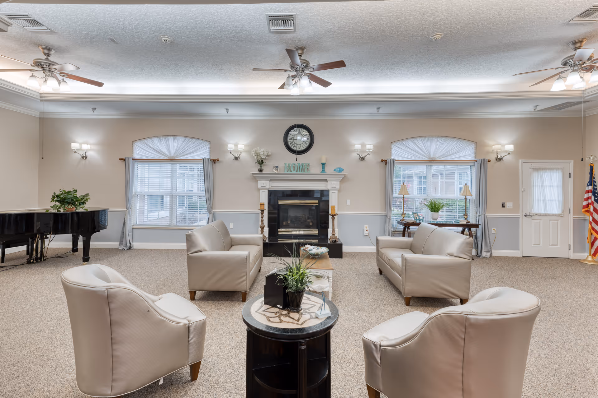 A spacious and well-lit living room with beige walls and carpeted floor. The room features two beige sofas and two beige armchairs arranged around a small round table with a plant on it. There is a fireplace with decorative items and a clock above it on the far wall, flanked by two windows with light blue curtains. A black grand piano is positioned on the left side of the room. Ceiling fans with lights hang from the ceiling, and an American flag stands near a door on the right side.
