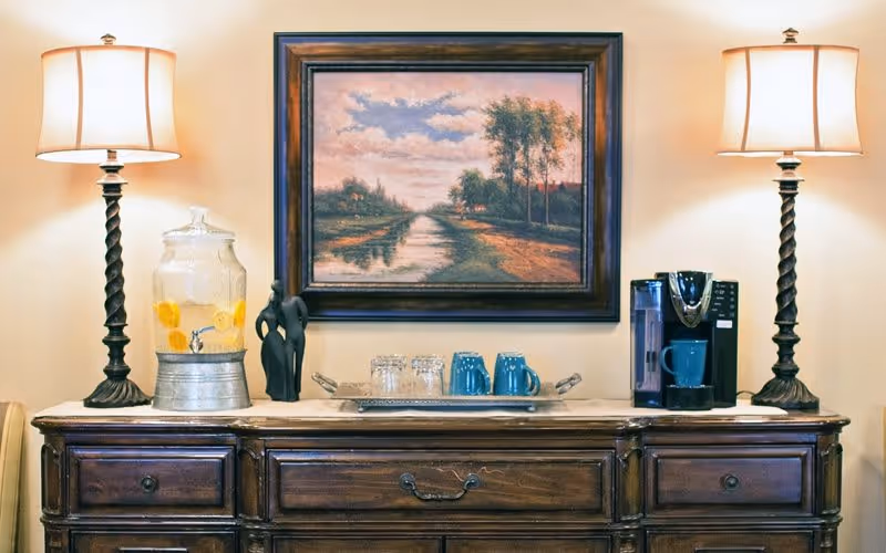 A wooden sideboard with two ornate table lamps on either end, a glass beverage dispenser with lemon water, a black abstract sculpture, a tray with blue mugs and glasses, and a coffee maker. Above the sideboard hangs a framed painting of a serene landscape with a river, trees, and a cloudy sky.