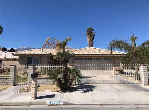 Front exterior of a single-story stucco house with a gated driveway, garage, and palm trees.