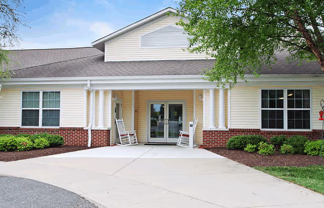 Entrance of a single-story senior living building with double glass doors, white columns, two rocking chairs, and surrounding landscaping.