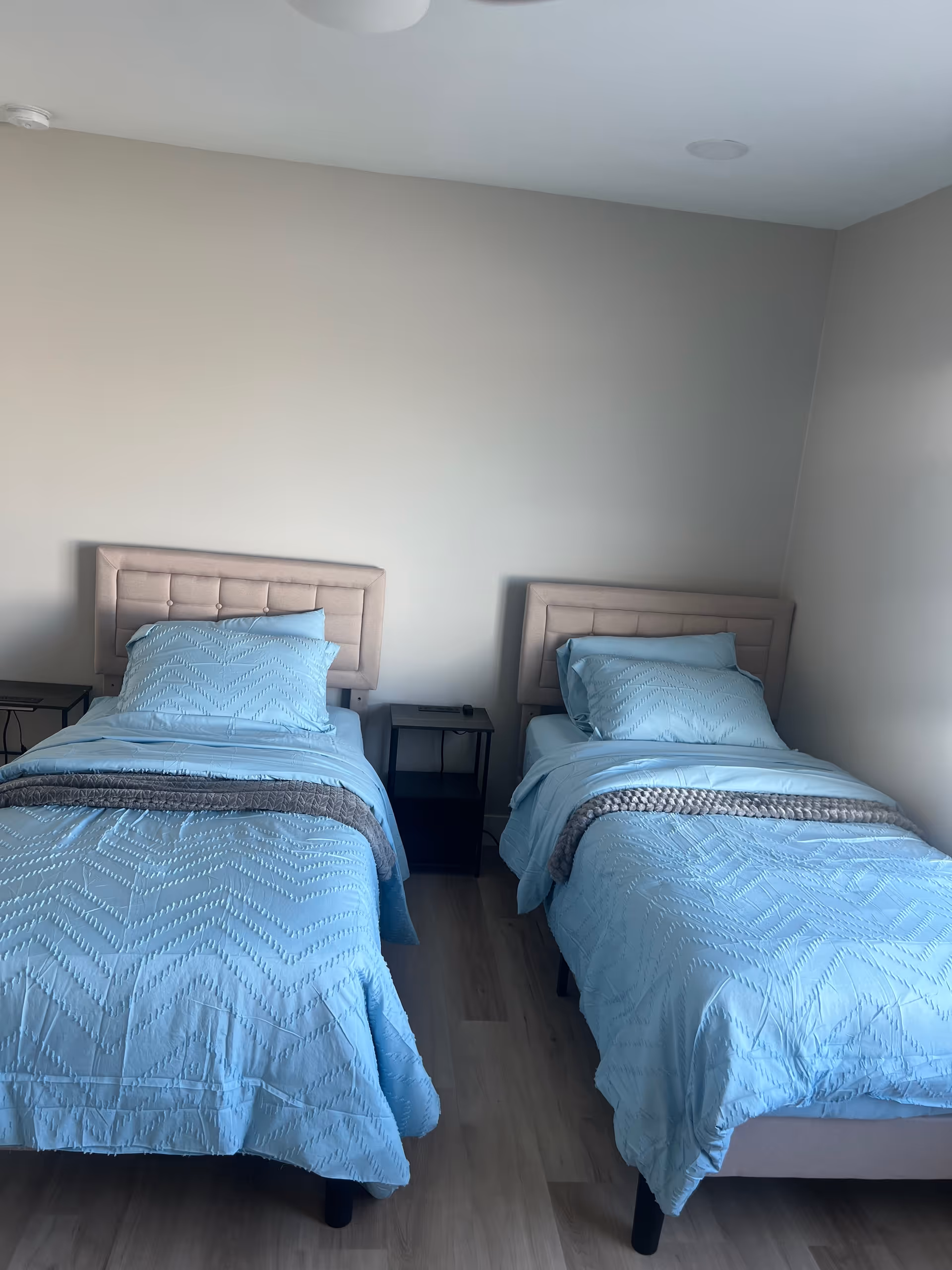 Two twin beds with light blue bedding and a small nightstand between them in a plain bedroom.