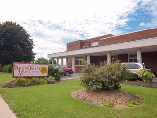 Exterior view of Saucon Valley Manor building with a covered entrance, parked cars, and landscaped bushes and grass under a partly cloudy sky.
