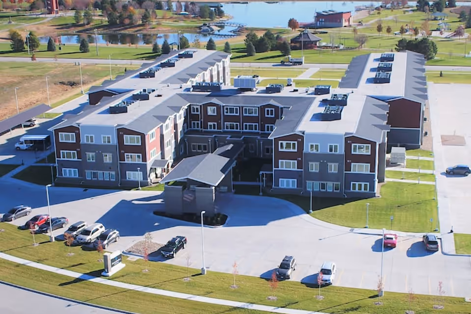 Aerial view of a large senior living facility building with multiple floors, surrounded by parking lots and green lawns. The building has a covered entrance and is situated near a body of water with trees and pathways in the background.
