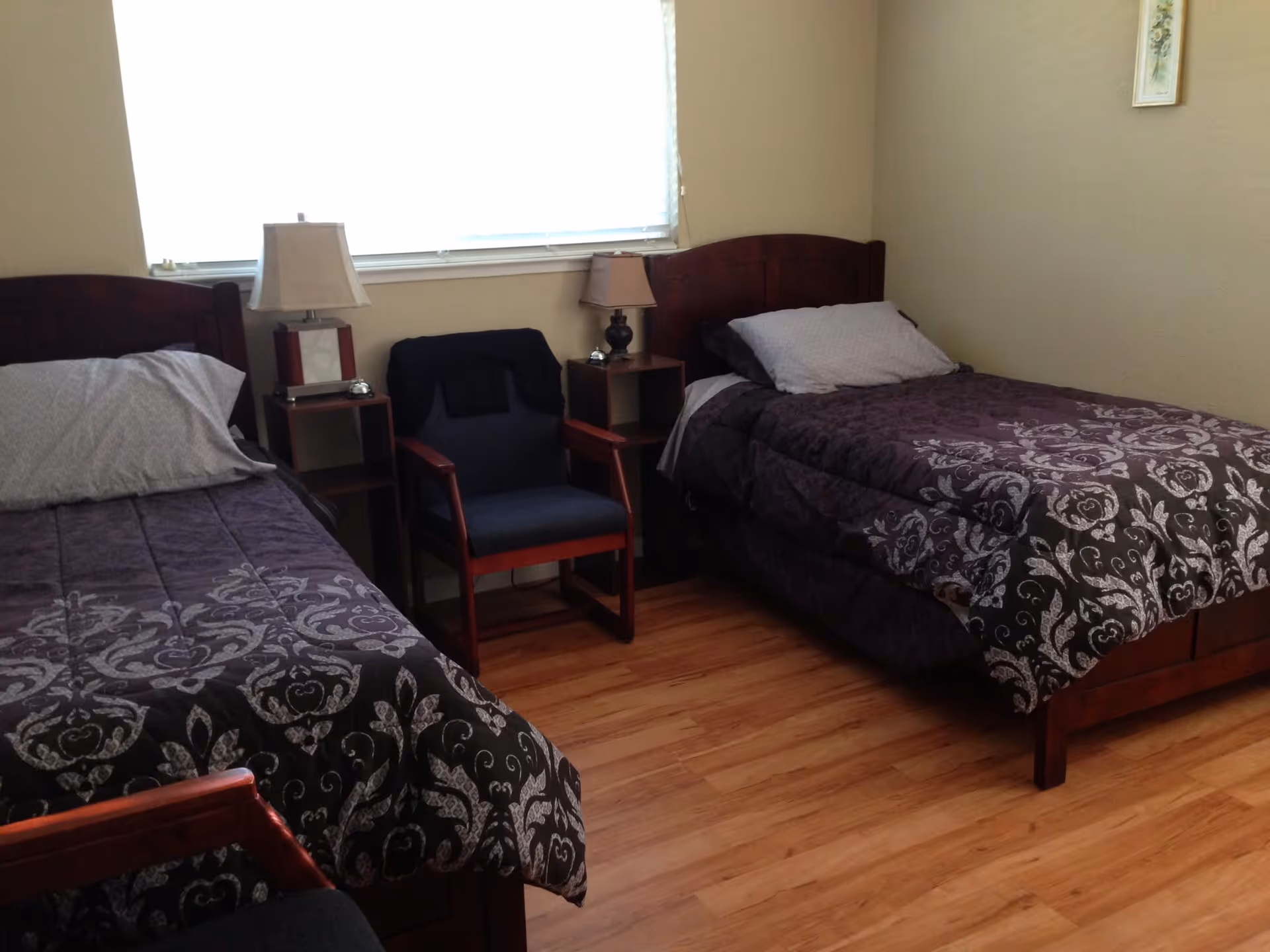 A tidy room with two twin beds with dark patterned comforters, a chair and bedside tables beneath a bright window.