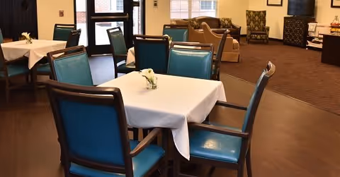 Interior view of a dining area with tables covered in white tablecloths and blue cushioned chairs. The room has large windows and doors letting in natural light, with additional seating and decor visible in the background.