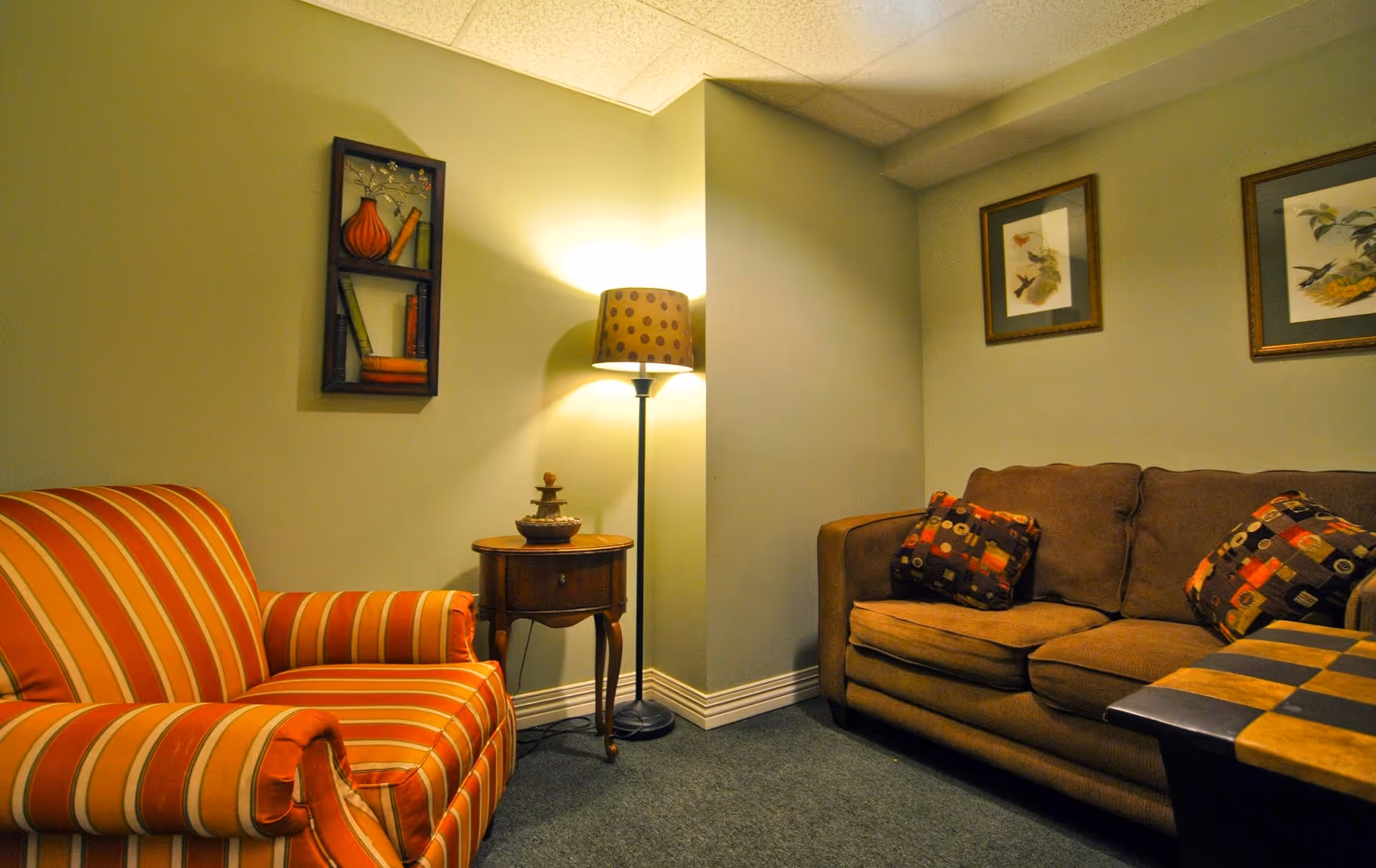 A cozy living room corner with a brown couch adorned with patterned pillows, a striped armchair, a small wooden side table with a decorative item, a floor lamp with a polka dot shade, and framed bird artwork on the walls.