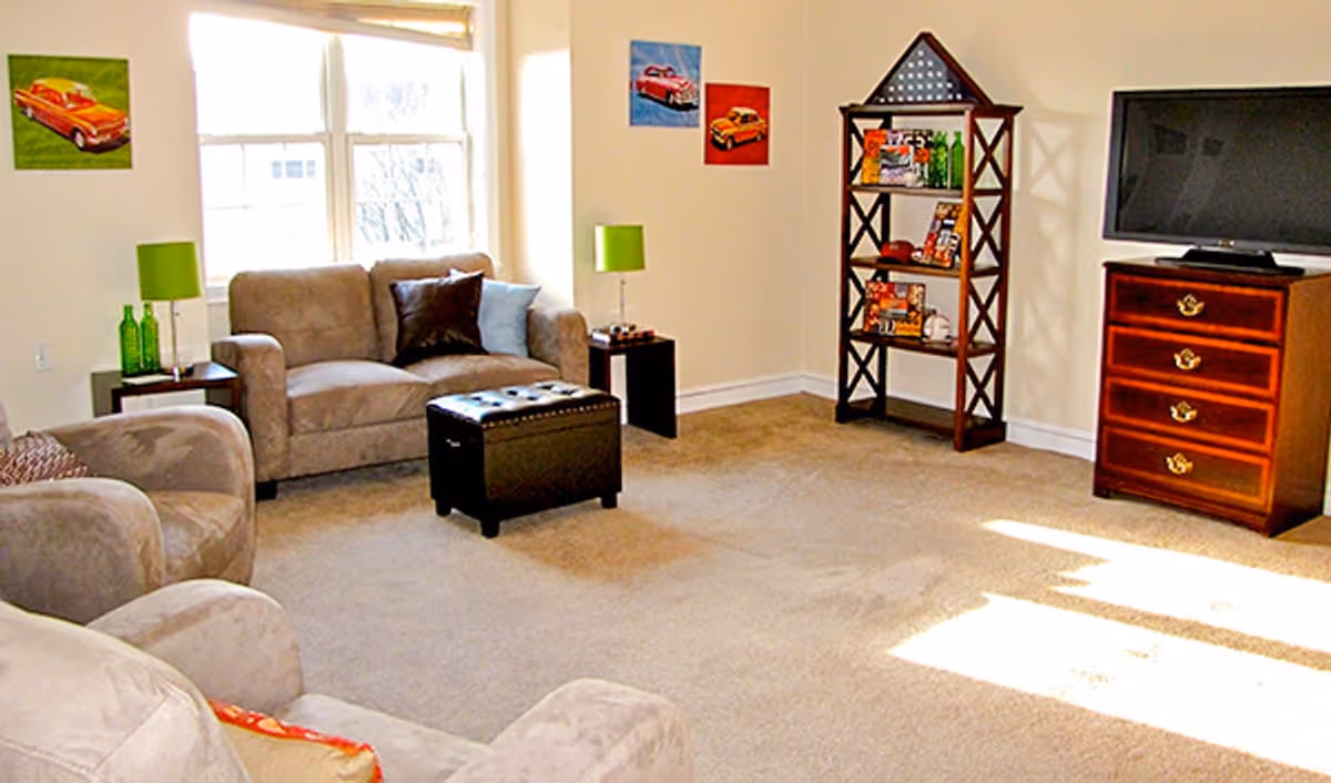 A bright living room with beige carpet and light-colored walls. The room features a beige loveseat with two pillows, a matching armchair, a black ottoman, two small side tables with green lamps, a wooden shelving unit with decorative items, and a wooden chest of drawers with a flat-screen TV on top. There are three small paintings of cars on the walls and a large window letting in natural light.