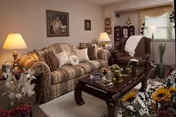 A cozy living room with a patterned sofa and armchair, a wooden coffee table with decorative items, table lamps, framed artwork on the walls, and various plants and flowers adding a warm and inviting atmosphere.