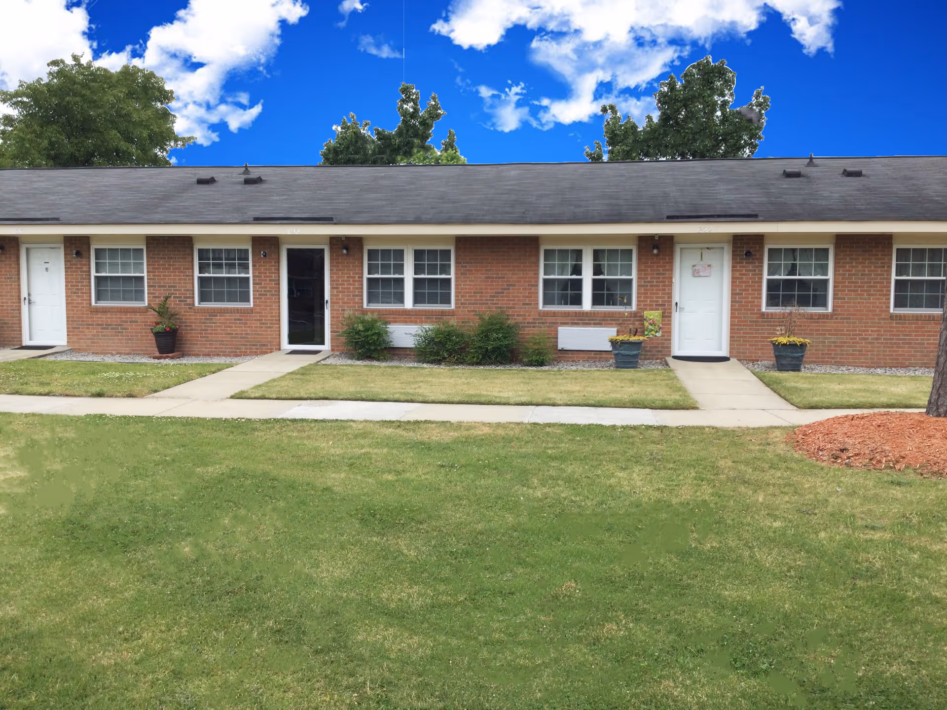 Exterior view of a single-story brick apartment building with multiple white doors and windows, a concrete walkway, green grass lawn, potted plants, and trees under a blue sky with scattered clouds.