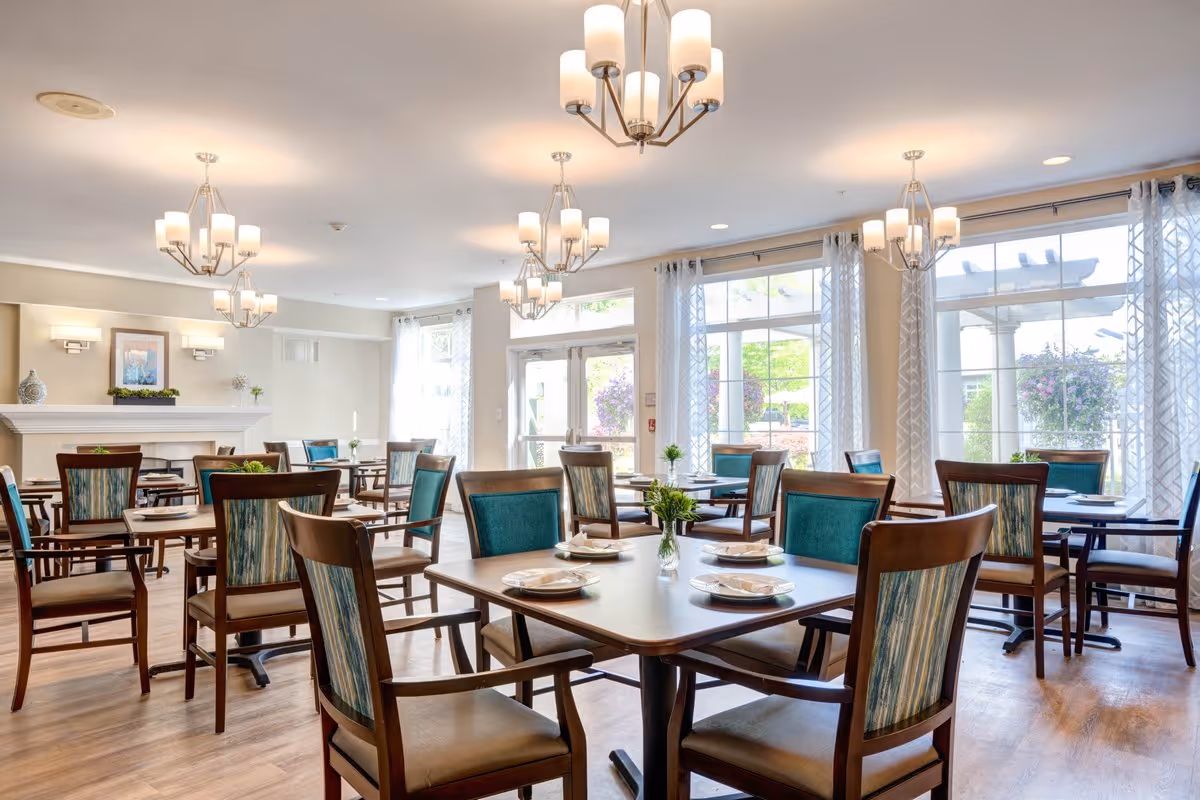 A bright dining room with multiple wooden tables and chairs featuring teal and patterned upholstery. Each table is set with plates and napkins, and small vases with green plants serve as centerpieces. Large windows with sheer curtains allow natural light to fill the room. Modern chandeliers hang from the ceiling, and a white fireplace with decorative items is visible in the background.