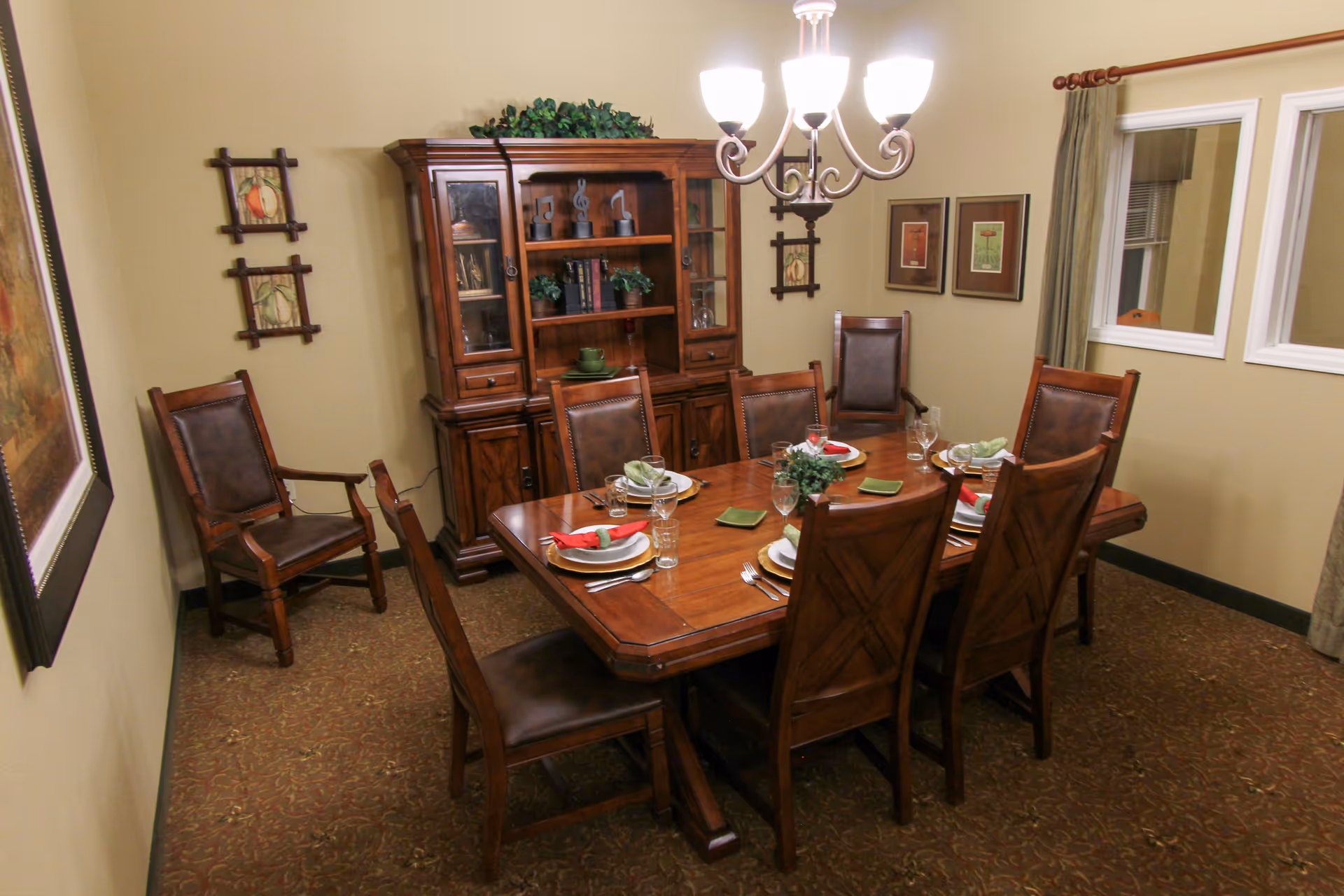 A dining room with a wooden dining table set for six people, featuring plates, glasses, napkins, and silverware. The room has six wooden chairs with leather cushions, a wooden cabinet with glass doors displaying decorative items, framed artwork on the walls, a chandelier overhead, and two windows with curtains.
