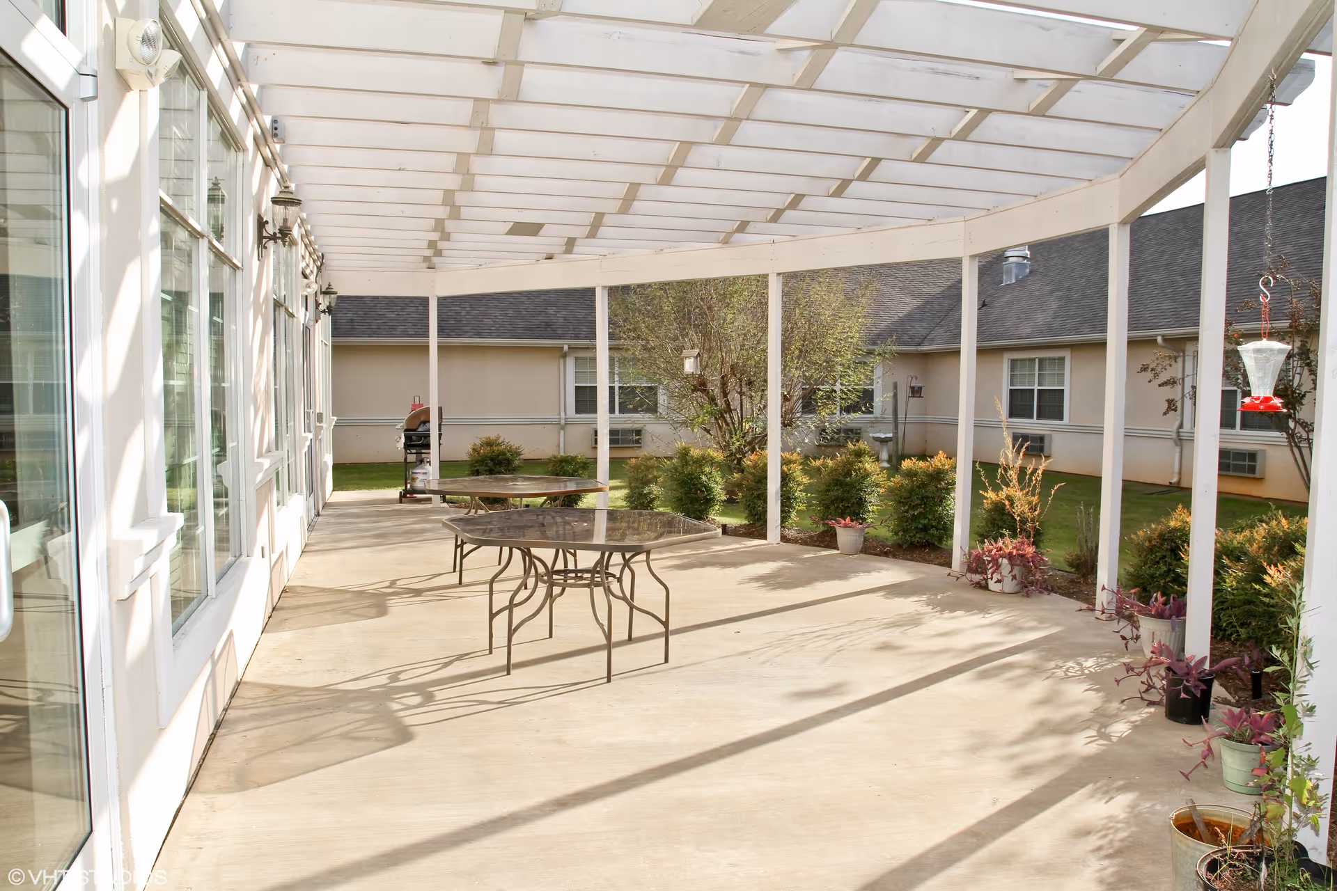 A covered outdoor patio area with a white pergola roof, two round glass-top tables with metal legs, potted plants along the edges, and a view of a garden and building windows in the background.