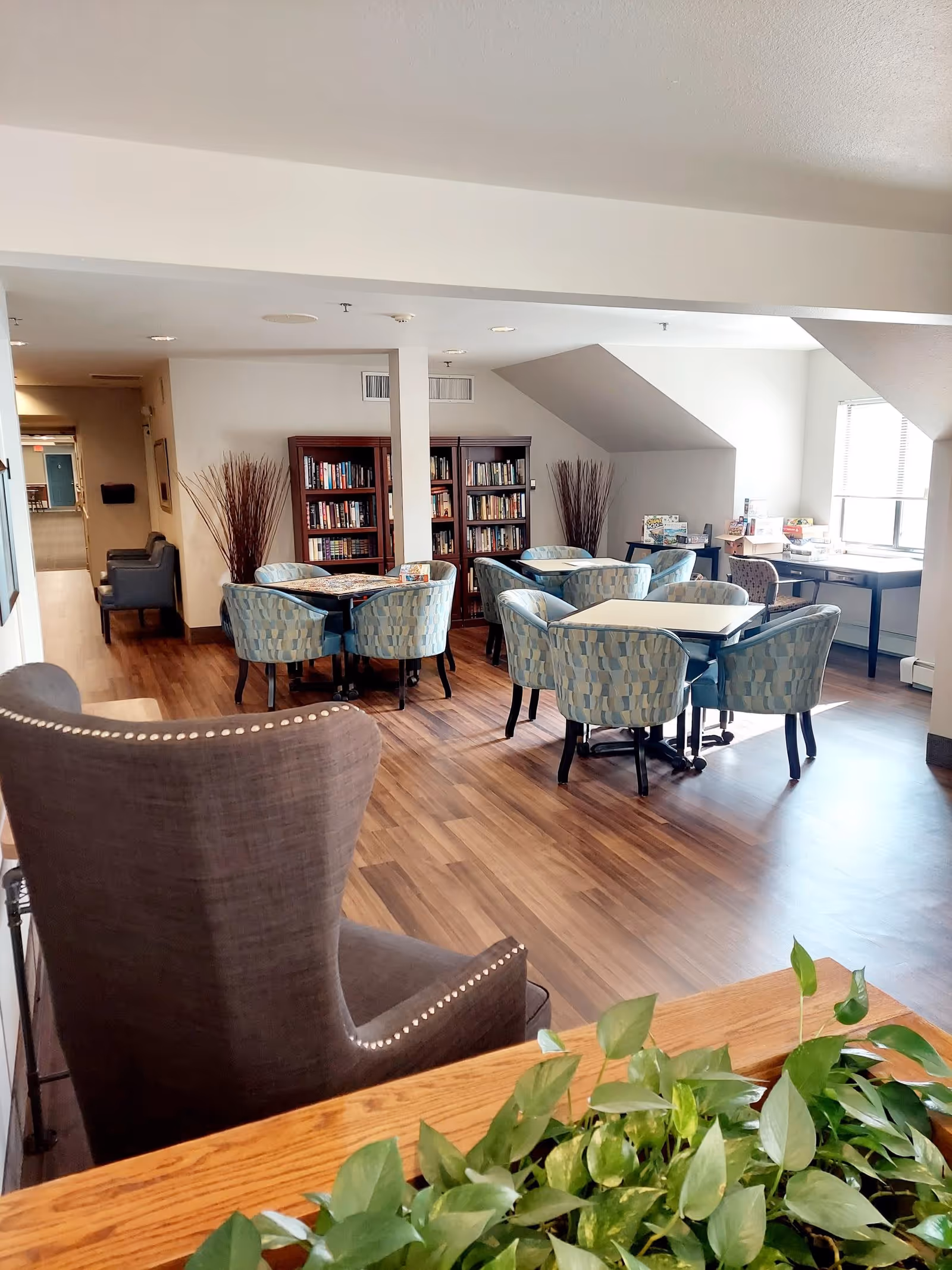 A bright common area in a senior living facility with wooden flooring, several tables surrounded by patterned blue chairs, two tall bookshelves filled with books, and a window letting in natural light. There is a large gray armchair in the foreground and some green plants on a wooden surface.