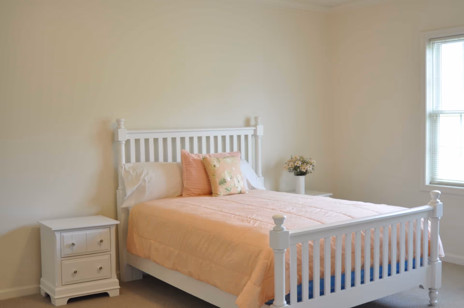 A bedroom with a white wooden bed frame, a peach-colored comforter, two pillows with floral and striped patterns, a white nightstand with two drawers on the left side, and a vase with flowers on a small table near a window with blinds.
