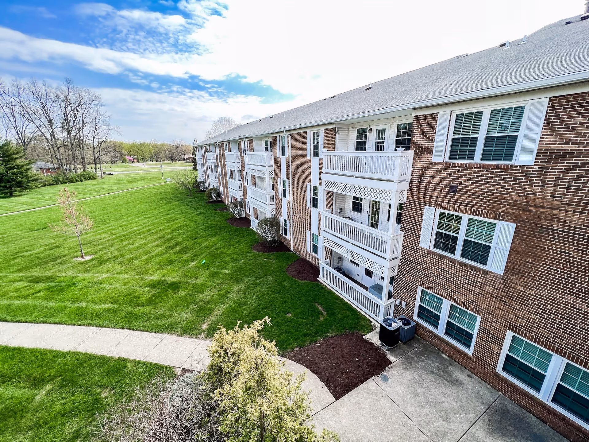 Exterior view of a multi-story brick residential building with white balconies and windows, surrounded by a well-maintained green lawn and a concrete walkway under a partly cloudy sky.