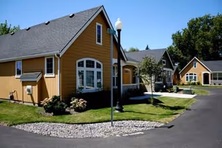 Exterior view of a senior living facility with multiple yellow and gray buildings, a paved driveway, green lawns, and trees under a clear blue sky.