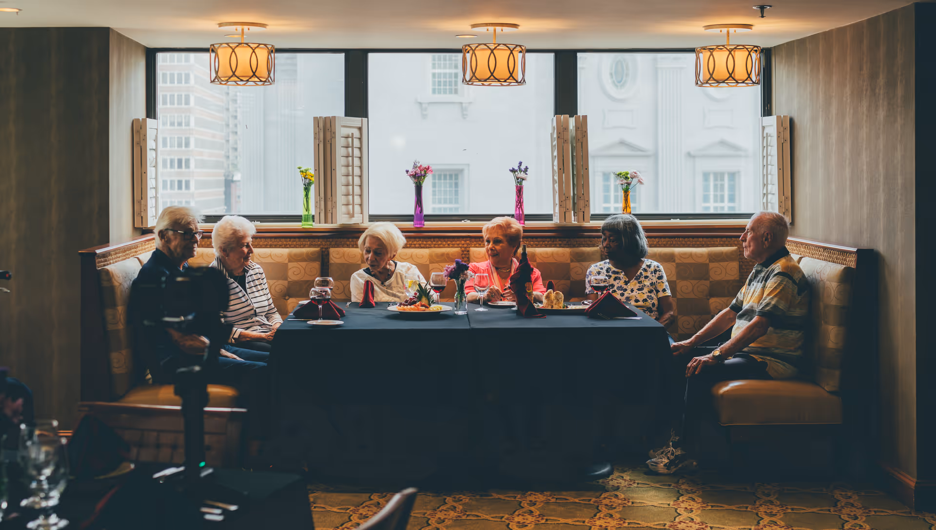 A group of six elderly residents sit around a table in a cozy, windowed dining nook with decorative lighting and vases.