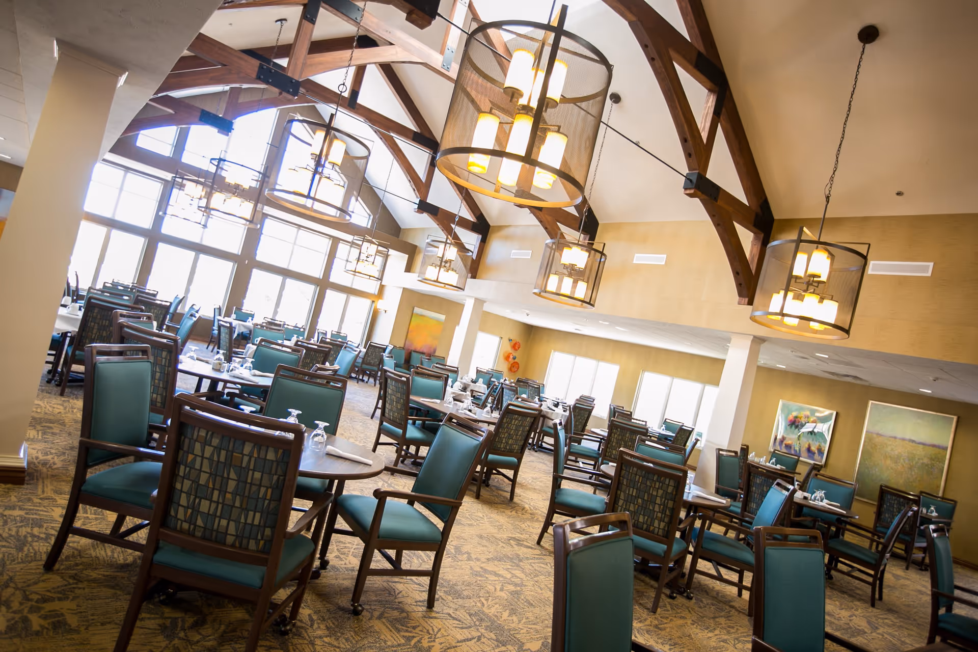 Spacious dining room with multiple tables and teal cushioned chairs arranged neatly. Large windows allow natural light to fill the room, and modern hanging light fixtures with wooden beams adorn the ceiling. The walls are decorated with colorful paintings.