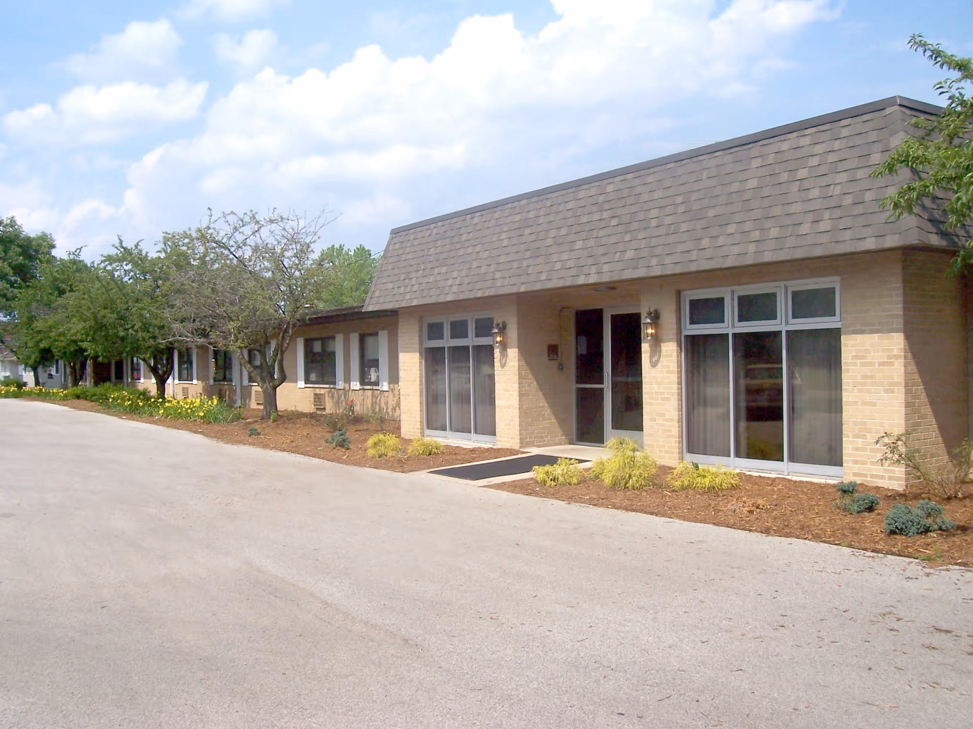 Low-rise brick building entrance with large windows, landscaping, and a paved driveway under a partly cloudy sky.