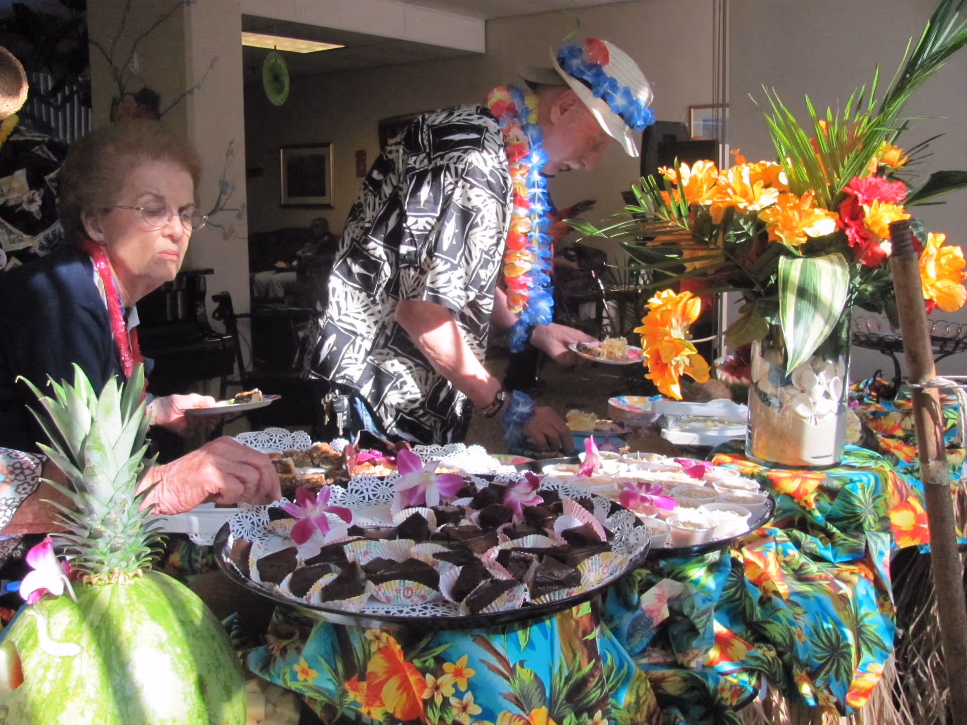 Two older adults serve themselves at a tropical-themed buffet table decorated with bright flowers and trays of desserts.