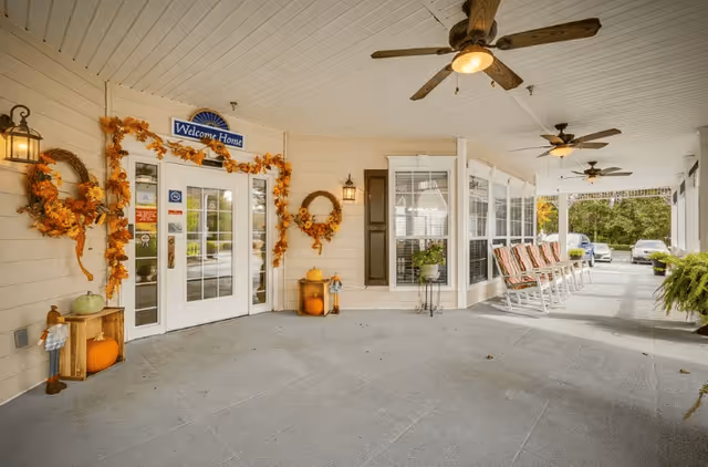 Covered entrance area of a senior living facility decorated with autumn-themed wreaths and garlands around the door. The door has a 'Welcome Home' sign above it. There are rocking chairs lined up along the right side under the covered porch, with ceiling fans overhead. Some pumpkins and decorative items are placed near the door. Cars are visible parked in the background.