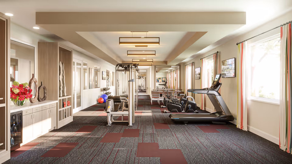 Bright, well-equipped fitness room with treadmills, weight machines, exercise balls, and seating along windows.