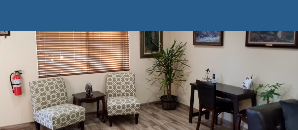 A cozy interior room with two patterned armchairs separated by a small wooden table, a potted plant in the corner, a dark wooden desk with a chair, and framed artwork on the walls. The floor has wood-like flooring and a window with wooden blinds is visible.