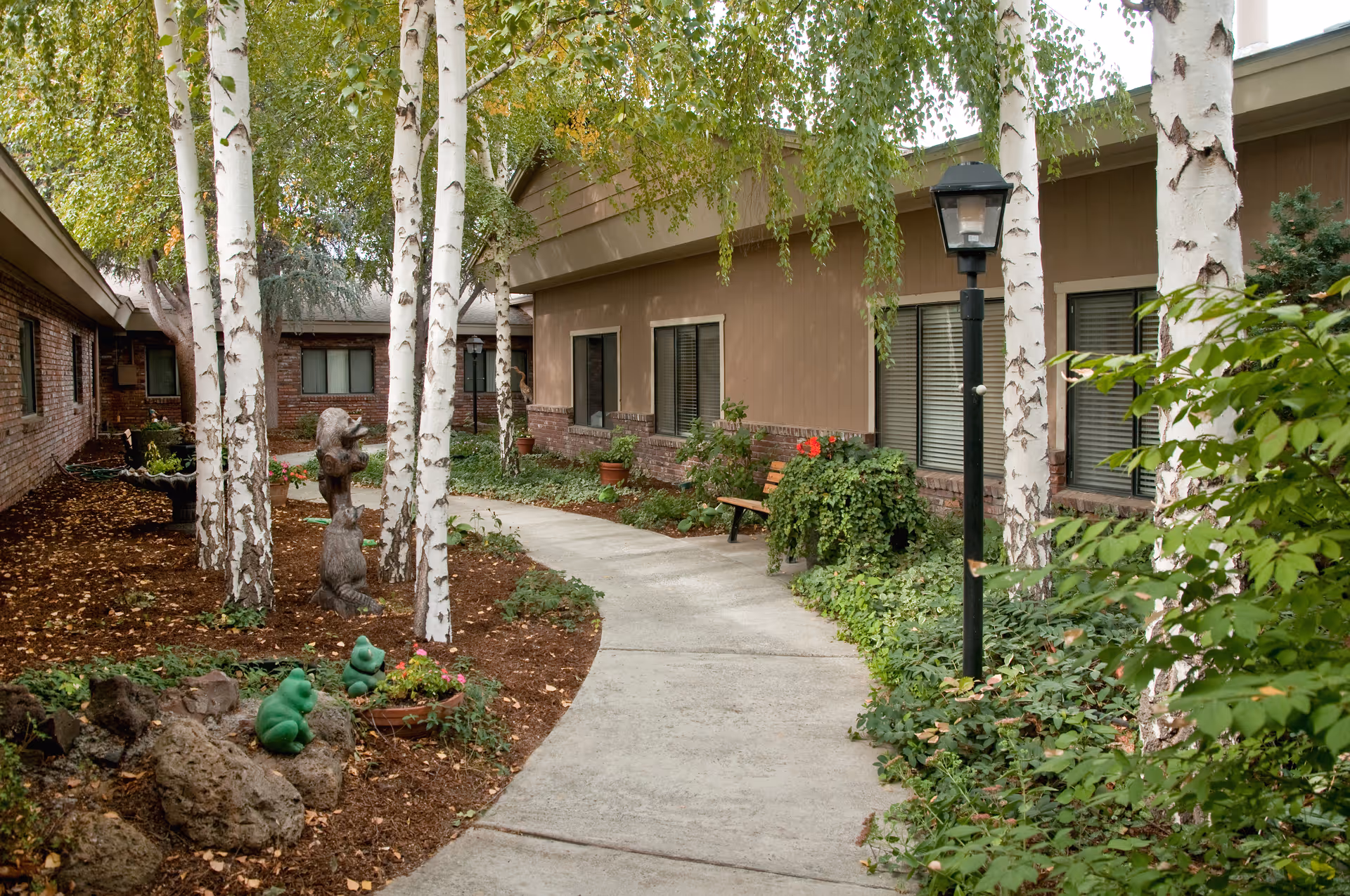 A peaceful outdoor courtyard area with a curved concrete walkway surrounded by trees, plants, and shrubs. There are decorative statues of bears and frogs, a wooden bench, and a lamp post. The courtyard is enclosed by a building with brick and beige siding and several windows.