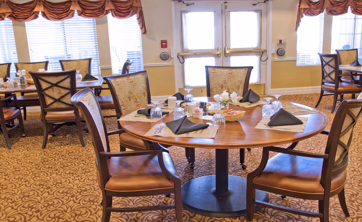 A dining room in a senior living facility with round wooden tables set for four people. Each place setting includes a placemat, black folded napkin, upside-down glass, and silverware. The chairs have cushioned seats and floral-patterned backs. The room has carpeted floors, large windows with blinds and valances, and double doors in the background.