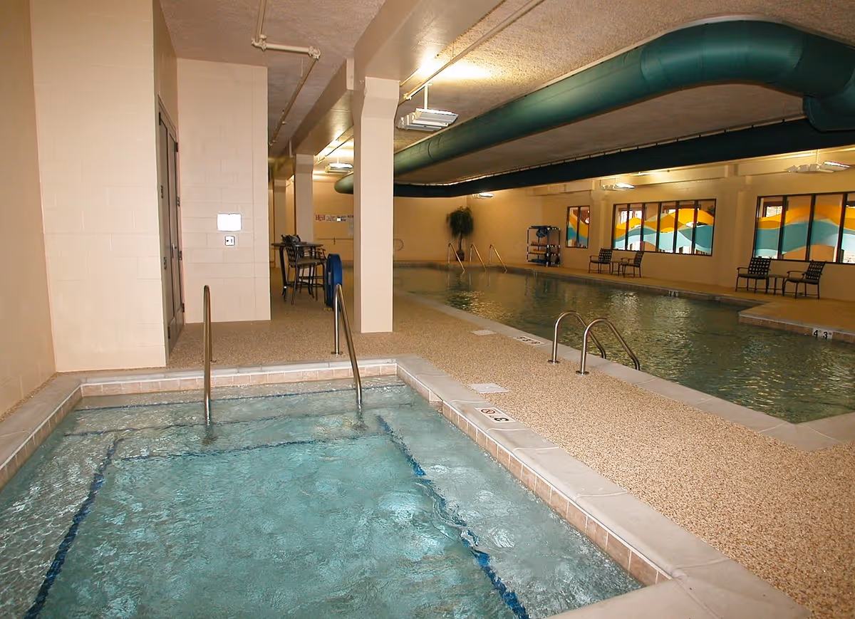 Indoor pool area with a hot tub in the foreground and a swimming pool in the background. The room has beige walls, a textured ceiling with exposed ductwork painted green, and several chairs and tables along the far wall with colorful wave-patterned windows.